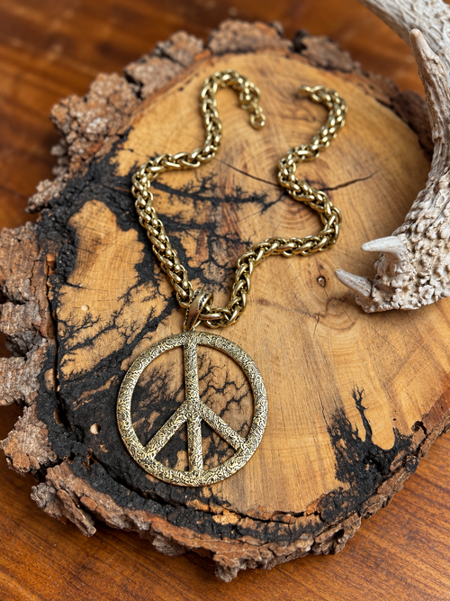 Antiqued gold-tone peace sign pendant with textured detailing and a chunky chain on rustic wood