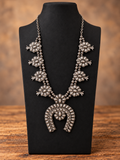 Silver-Tone Floral Squash Blossom Necklace displayed on a jewelry stand against a warm wood tabletop and softly blurred brown background