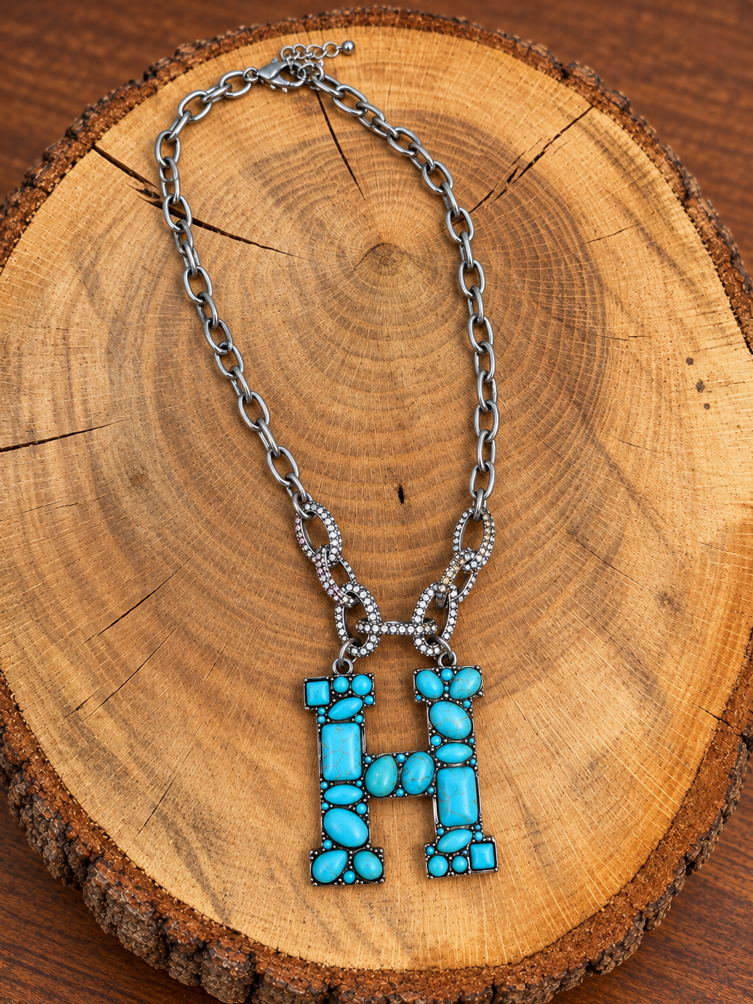 Turquoise letter H pendant with rhinestone accents on a silver-tone chain, displayed on warm rustic wood