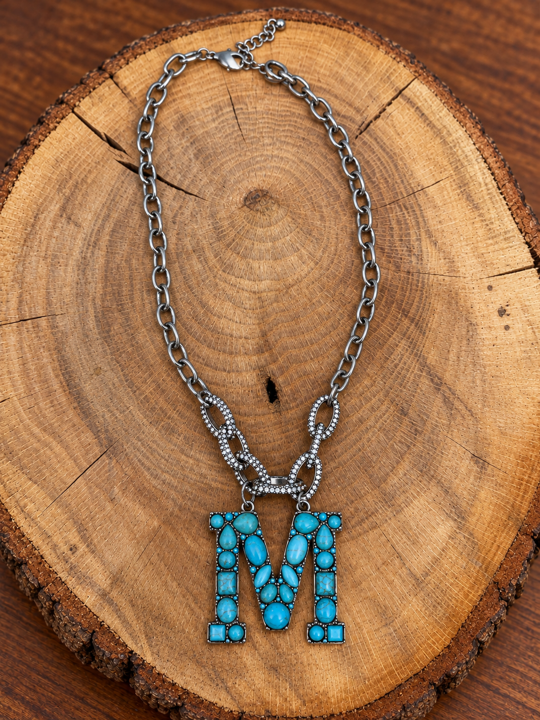 Turquoise letter M pendant with rhinestone accents on a silver-tone chain, displayed on warm rustic wood