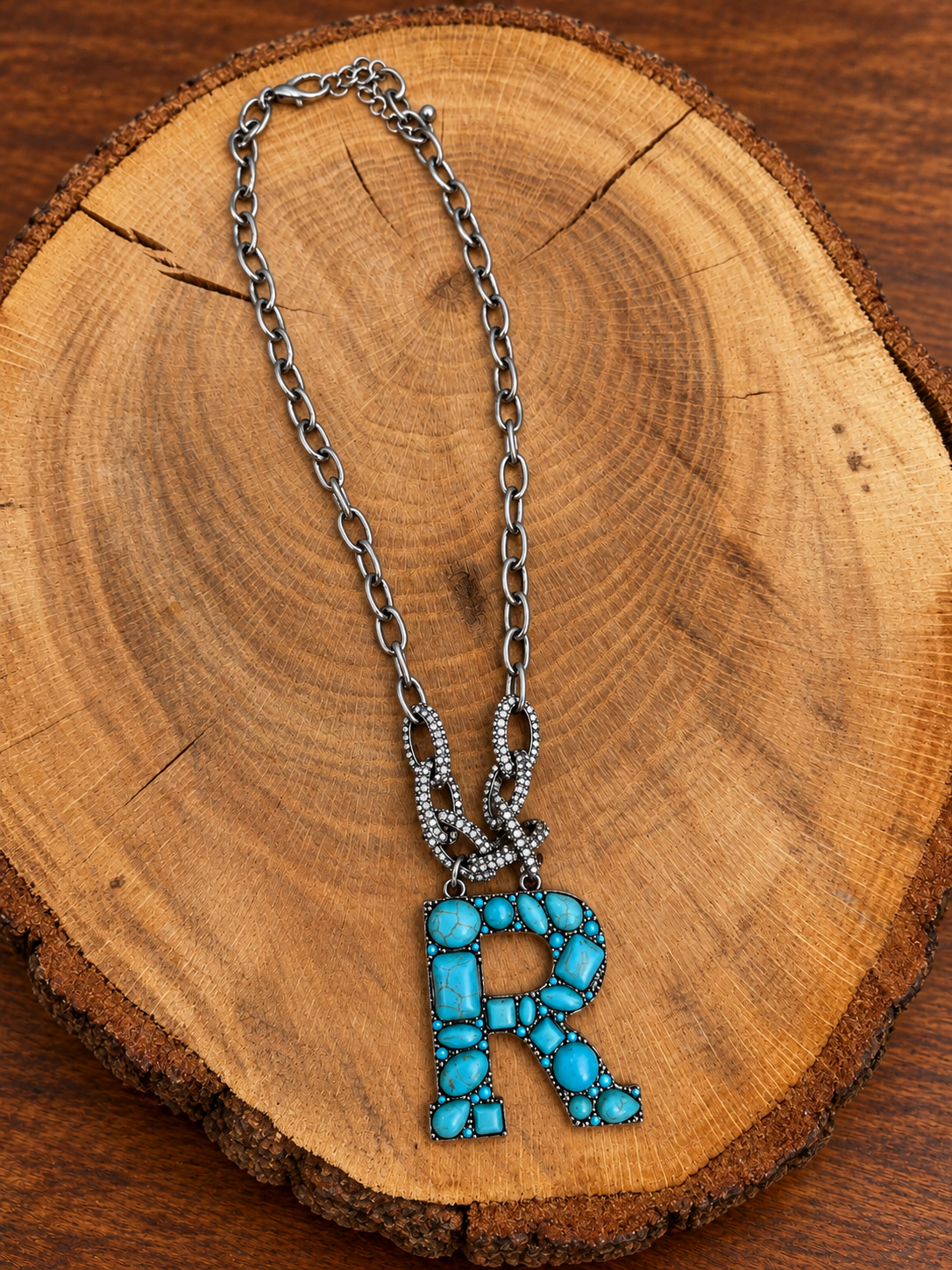 Turquoise letter R pendant with rhinestone accents on a silver-tone chain, displayed on warm rustic wood